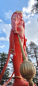 A large statue of a mythological figure with pinkish-red clothing and a golden crown, holding a mace. The statue is surrounded by trees under a blue sky with scattered clouds.