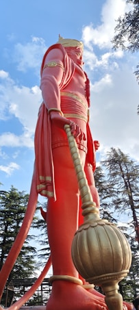 A large statue of a mythological figure with pinkish-red clothing and a golden crown, holding a mace. The statue is surrounded by trees under a blue sky with scattered clouds.