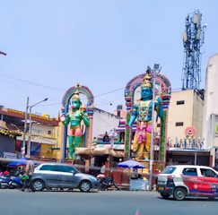 Two large, colorful statues depicting Hindu deities stand prominently in a busy urban setting. The statues are adorned with intricate details and vibrant colors, with one holding a weapon and the other in a standing posture. The scene includes surrounding buildings, vehicles, and a tower in the background. The environment has an outdoor market or a temple atmosphere.