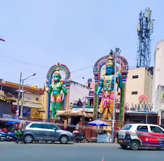 Two large, colorful statues depicting Hindu deities stand prominently in a busy urban setting. The statues are adorned with intricate details and vibrant colors, with one holding a weapon and the other in a standing posture. The scene includes surrounding buildings, vehicles, and a tower in the background. The environment has an outdoor market or a temple atmosphere.