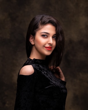 A woman with long dark hair wearing a black velvet top with shoulder cutouts. Her makeup is elegant and her lips are painted a vibrant red. The background is a dark, textured backdrop, which contrasts with her smooth skin and the black attire.
