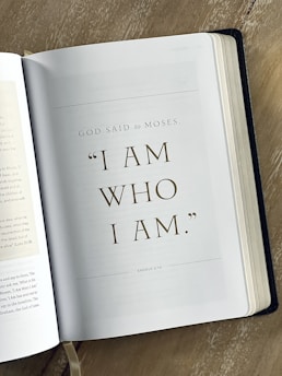 An open book displaying a page with a biblical quote from Exodus 3:14, 'I AM WHO I AM,' attributed to God speaking to Moses. The text is centered on the page in large, elegant typography. The book rests on a wooden surface, with the left-hand page slightly visible.