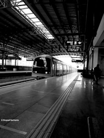 A black and white image of a train station with a train on the platform. The station features a high, geometric roof structure and a shiny floor with guiding tracks. There is signage visible above the platform indicating directions. A silhouette of a person stands to the right side of the image.