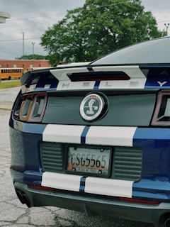 A close-up of the rear end of a blue car with white racing stripes. The license plate indicates it's registered in Georgia. The emblem reads 'Shelby GT500' with the iconic cobra logo. Trees and a yellow school bus are visible in the background.