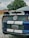 A close-up of the rear end of a blue car with white racing stripes. The license plate indicates it's registered in Georgia. The emblem reads 'Shelby GT500' with the iconic cobra logo. Trees and a yellow school bus are visible in the background.