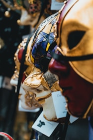 Elegant Venetian masks are displayed in close-up, showcasing intricate designs and rich colors. The masks feature gold detailing, vivid patterns, and textures that evoke a sense of mystery and artistry.
