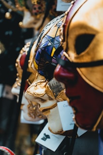 Elegant Venetian masks are displayed in close-up, showcasing intricate designs and rich colors. The masks feature gold detailing, vivid patterns, and textures that evoke a sense of mystery and artistry.
