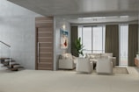 A modern living room with a minimalist design. The room features a comfortable beige sectional sofa and two matching armchairs on a neutral area rug. A sleek wooden door is adjacent to a contemporary staircase with glass railings. Floor-to-ceiling windows with sheer and heavy curtains offer a view outside. A potted plant adds a touch of greenery next to the sofa, and a colorful painting adorns the wall.