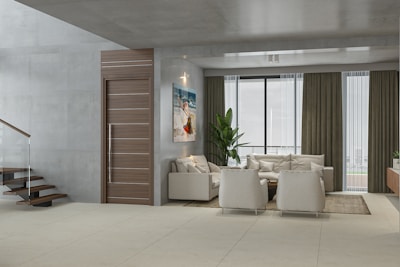 A modern living room with a minimalist design. The room features a comfortable beige sectional sofa and two matching armchairs on a neutral area rug. A sleek wooden door is adjacent to a contemporary staircase with glass railings. Floor-to-ceiling windows with sheer and heavy curtains offer a view outside. A potted plant adds a touch of greenery next to the sofa, and a colorful painting adorns the wall.