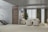 A modern living room with a minimalist design. The room features a comfortable beige sectional sofa and two matching armchairs on a neutral area rug. A sleek wooden door is adjacent to a contemporary staircase with glass railings. Floor-to-ceiling windows with sheer and heavy curtains offer a view outside. A potted plant adds a touch of greenery next to the sofa, and a colorful painting adorns the wall.