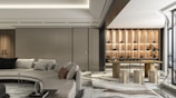 A modern interior space featuring a large beige sectional sofa with several cushions, adjacent to a wooden table with stools. The flooring has a glossy marble finish, and there is an elegant wall with shelves lit to display decorative items. A piece of modern art or a bonsai tree enhances the room's contemporary design. Large windows allow natural light to fill the open space, enhancing its luxurious atmosphere.