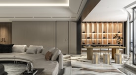 A modern interior space featuring a large beige sectional sofa with several cushions, adjacent to a wooden table with stools. The flooring has a glossy marble finish, and there is an elegant wall with shelves lit to display decorative items. A piece of modern art or a bonsai tree enhances the room's contemporary design. Large windows allow natural light to fill the open space, enhancing its luxurious atmosphere.