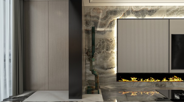 A modern, sleek interior with a vertical, ribbed panel integrated into a decorative wall. Beneath the panel, a linear gas fireplace emits artificial flames. The wall features a marble texture with a mix of dark and light tones. To the left, a section of softly pleated gray curtains is visible, adding an element of elegance.