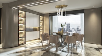 A modern dining area features a glass table surrounded by six beige chairs. The room is well-lit by both natural light from a large window with cityscape views and ambient lighting fixtures. The flooring is polished with a marble pattern, and the walls have a sleek, wood finish with built-in shelving units housing bottles.