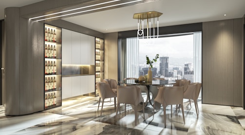 A modern dining area features a glass table surrounded by six beige chairs. The room is well-lit by both natural light from a large window with cityscape views and ambient lighting fixtures. The flooring is polished with a marble pattern, and the walls have a sleek, wood finish with built-in shelving units housing bottles.