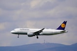 A commercial airplane in mid-flight with a clear view of its branding, flying against a background of cloudy skies and distant hills.