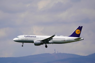 A commercial airplane in mid-flight with a clear view of its branding, flying against a background of cloudy skies and distant hills.