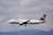 A commercial airplane in mid-flight with a clear view of its branding, flying against a background of cloudy skies and distant hills.