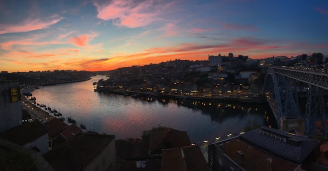 A picturesque view of a cityscape at sunset with a river running through the middle. The sky is filled with colorful hues of pink, orange, and blue as the sun sets. A large bridge spans the river on the right, and the city is bustling with lights that reflect in the water. Buildings with terracotta roofs are visible in the foreground, leading to a densely packed urban area across the river.