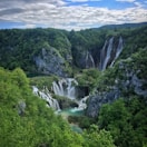 A stunning natural landscape featuring multiple cascading waterfalls surrounded by lush green forests and rocky cliffs under a partly cloudy sky. The water appears clear and turquoise, adding to the pristine beauty of the scene.