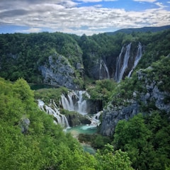 A stunning natural landscape featuring multiple cascading waterfalls surrounded by lush green forests and rocky cliffs under a partly cloudy sky. The water appears clear and turquoise, adding to the pristine beauty of the scene.
