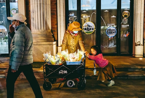 An elderly person is wearing a brown hat and face mask while pushing a cart filled with colorful bouquets. A small child in a bright pink top holds onto the cart, gazing in a different direction. The setting appears to be an urban street, potentially a shopping area, with storefront windows in the background displaying graphics and text.