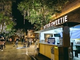 A food truck named 'Bitter Sweetie' is parked under illuminated tree branches decorated with hanging lights. People are gathered around in the open area, with some walking and others looking at their surroundings. The scene is set at night, creating a lively atmosphere. The food truck has a yellow exterior with lit signage and a punchy quote displayed on it. To the side, there are payment machines attached.