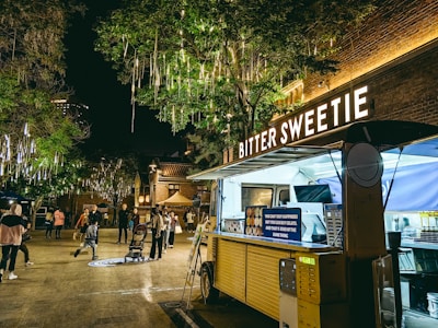 A food truck named 'Bitter Sweetie' is parked under illuminated tree branches decorated with hanging lights. People are gathered around in the open area, with some walking and others looking at their surroundings. The scene is set at night, creating a lively atmosphere. The food truck has a yellow exterior with lit signage and a punchy quote displayed on it. To the side, there are payment machines attached.