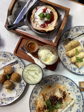 A cozy kitchen table displaying a variety of homemade dishes including malla, pastries, and spring rolls arranged on rustic plates.