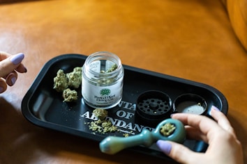 A tabletop scene with cannabis buds, a grinder, a labeled jar reading 'Perfect Plant Hemp Co,' and a person handling a green pipe. The setting is indoors on a brown surface.