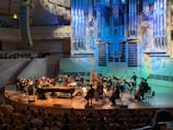 An orchestra is performing on stage with a conductor leading the musicians. The stage is illuminated with blue and yellow lighting, highlighting various musical instruments including pianos, string instruments, and a harp. The audience is seated and watching the performance in a large concert hall with a prominent pipe organ situated in the back of the stage. The venue has wood paneling and several tiers for seating.