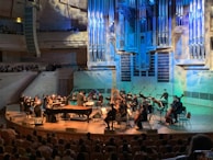 An orchestra is performing on stage with a conductor leading the musicians. The stage is illuminated with blue and yellow lighting, highlighting various musical instruments including pianos, string instruments, and a harp. The audience is seated and watching the performance in a large concert hall with a prominent pipe organ situated in the back of the stage. The venue has wood paneling and several tiers for seating.