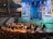 An orchestra is performing on stage with a conductor leading the musicians. The stage is illuminated with blue and yellow lighting, highlighting various musical instruments including pianos, string instruments, and a harp. The audience is seated and watching the performance in a large concert hall with a prominent pipe organ situated in the back of the stage. The venue has wood paneling and several tiers for seating.