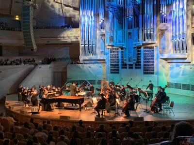 An orchestra is performing on stage with a conductor leading the musicians. The stage is illuminated with blue and yellow lighting, highlighting various musical instruments including pianos, string instruments, and a harp. The audience is seated and watching the performance in a large concert hall with a prominent pipe organ situated in the back of the stage. The venue has wood paneling and several tiers for seating.