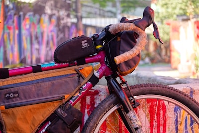 A close-up of a well-equipped bicycle is parked next to a colorful, graffiti-covered wall. The bike has several attached bags, including one on the frame and another on the handlebars. The handlebar has tape wrapped around it for grip, and the tire is a rugged, off-road type.
