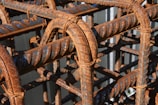 Rusted metal rebars intricately woven together, creating a grid-like structure. The metal displays signs of corrosion with brown rust overtaking the originally blue-gray surface. Sunlight casts shadows creating patterns and emphasizing the textured spirals on the bars.
