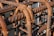 Rusted metal rebars intricately woven together, creating a grid-like structure. The metal displays signs of corrosion with brown rust overtaking the originally blue-gray surface. Sunlight casts shadows creating patterns and emphasizing the textured spirals on the bars.