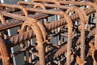 A close-up view of a grid structure made from rusted steel rebar, with each bar intricately twisted and held together by looping wire ties. The rusty texture and patina are clearly visible, indicating weathering and age.