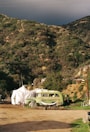 An old green bus is parked next to a white tent in a rural setting. The area is surrounded by lush, green hills under a moody sky. Various small items and outdoor furniture are scattered around, suggesting a makeshift campsite or living space.