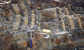 A rocky surface with layered sedimentary formations is visible. A geological hammer with a blue handle and a yellow measuring block are placed on the rocks, likely indicating a site of geological study or exploration.
