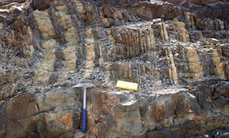 A rocky surface with layered sedimentary formations is visible. A geological hammer with a blue handle and a yellow measuring block are placed on the rocks, likely indicating a site of geological study or exploration.