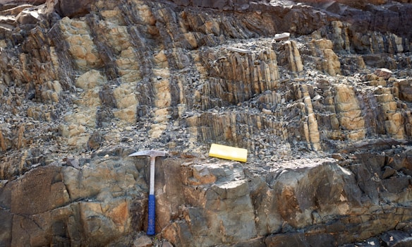 A rocky surface with layered sedimentary formations is visible. A geological hammer with a blue handle and a yellow measuring block are placed on the rocks, likely indicating a site of geological study or exploration.