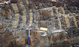 A rocky surface with layered sedimentary formations is visible. A geological hammer with a blue handle and a yellow measuring block are placed on the rocks, likely indicating a site of geological study or exploration.