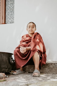 A young person wearing a maroon robe sits calmly on a stone surface. They have short hair and are wearing sandals. The background is a plain white wall with a small section of a window visible, featuring a decorative metal grill.