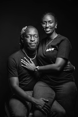 A couple is sitting together in an intimate pose, both wearing casual clothing and glasses. The woman is seated on the man's lap, and they are both smiling warmly. The setting is monochromatic with a focus on their expressions and body language.
