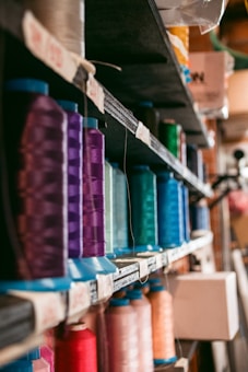 Rows of colorful thread spools neatly arranged on open shelves, each spool displaying a range of vibrant colors from purple and blue to red and green. The spools are organized by color and size, creating a visually appealing and organized appearance.
