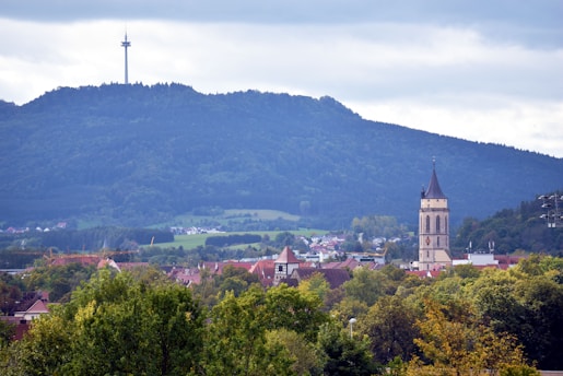 A picturesque landscape featuring a lush green hillside topped with a tall communications tower. In the foreground, there is a charming town with traditional buildings and a prominent church tower. The sky is overcast, adding a serene ambiance to the scene.