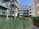 Modern apartment complex with large glass balconies and wooden paneling. The courtyard features lush greenery with ornamental grasses and young trees against a clear blue sky.