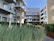 Modern apartment complex with large glass balconies and wooden paneling. The courtyard features lush greenery with ornamental grasses and young trees against a clear blue sky.