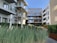 Modern apartment complex with large glass balconies and wooden paneling. The courtyard features lush greenery with ornamental grasses and young trees against a clear blue sky.